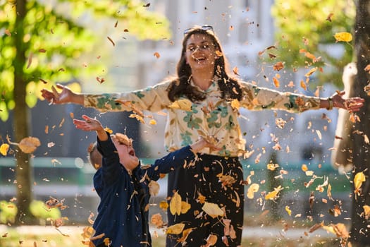 A modern woman joyfully plays with her son in the park, tossing leaves on a beautiful autumn day, capturing the essence of family life and the warmth of mother-son bonding in the midst of the fall season.