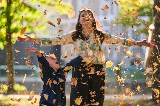 A modern woman joyfully plays with her son in the park, tossing leaves on a beautiful autumn day, capturing the essence of family life and the warmth of mother-son bonding in the midst of the fall season.