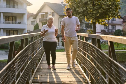 A handsome man and an older woman share a serene walk in nature, crossing a beautiful bridge against the backdrop of a stunning sunset, embodying the concept of a healthy and vibrant intergenerational life