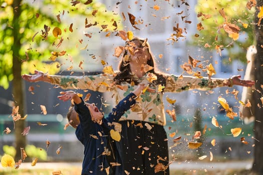 A modern woman joyfully plays with her son in the park, tossing leaves on a beautiful autumn day, capturing the essence of family life and the warmth of mother-son bonding in the midst of the fall season.