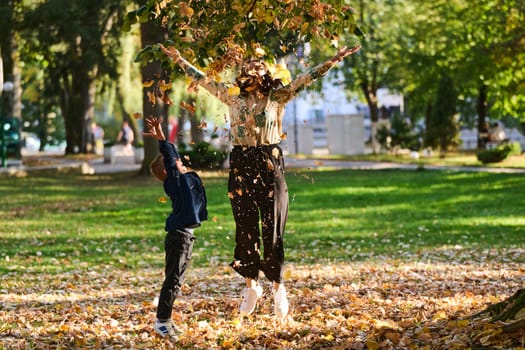 A modern woman joyfully plays with her son in the park, tossing leaves on a beautiful autumn day, capturing the essence of family life and the warmth of mother-son bonding in the midst of the fall season.