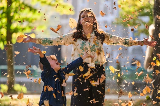 A modern woman joyfully plays with her son in the park, tossing leaves on a beautiful autumn day, capturing the essence of family life and the warmth of mother-son bonding in the midst of the fall season.