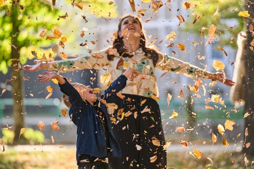 A modern woman joyfully plays with her son in the park, tossing leaves on a beautiful autumn day, capturing the essence of family life and the warmth of mother-son bonding in the midst of the fall season.