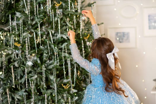 The girl decorates the Christmas tree, holding Christmas baubles in her hand. She wears a blue dress with a bow in her hair. Holiday concept