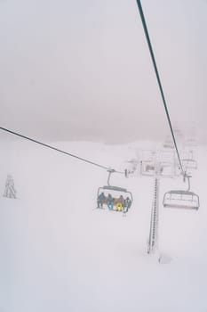 Tourists in colorful ski suits ride uphill through the fog on a chairlift above a snowy forest. High quality photo