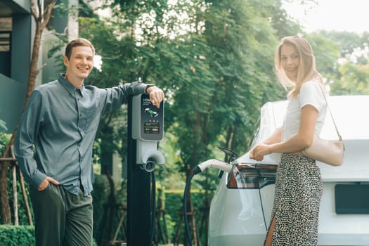 Young couple travel with EV electric car charging in green sustainable city outdoor garden in summer shows urban sustainability lifestyle by green clean rechargeable energy of electric vehicle innards