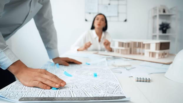 Skilled caucasian interior designer working with house model while discussing with asian architect engineer about house plan on meeting table with blueprint and house model scatter around. Immaculate.