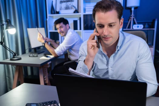 Serious businessman calling on phone with customer to create new project at night time, coworker writing document work sheet on desk. Concept of working on neon blue light modern office. Sellable.