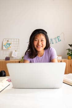Happy, smiling teen asian female high school student studying at home, doing homework using laptop looking at camera. Vertical image. Education, e-learning concept.