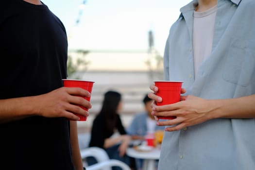 Two young male friends holding plastic glasses with beer at outdoor rooftop party.