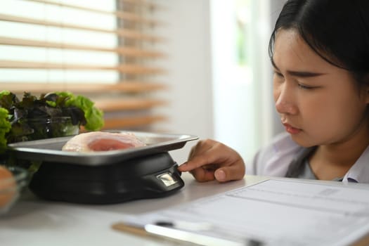 Female dietitian weighing raw chicken breast on a scale while working on a diet plan at clinic.