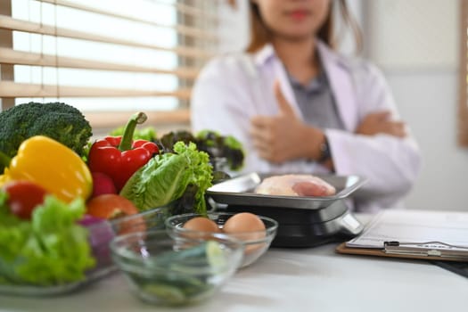 Closeup variety fresh vegetables, meat and eggs on white table with nutritionist sitting on background.