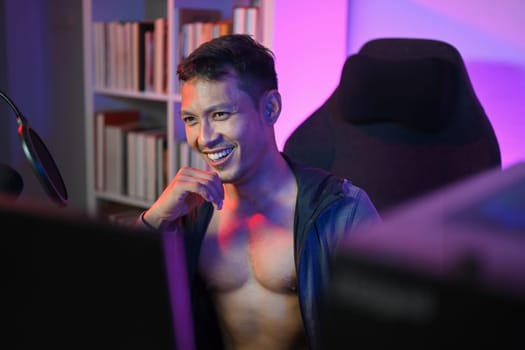 Confident muscled Asian male podcaster streaming audio podcast at home with neon lights.