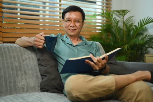 Positive middle age man in glasses drinking hot coffee and reading book on couch at home.