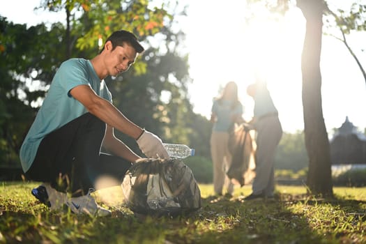 Smiling Asian male volunteer and his colleagues picking up plastic trash in public park.