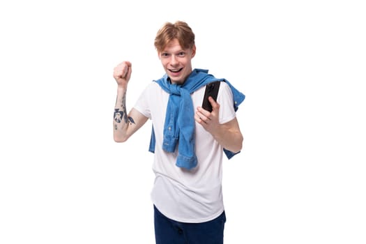 young fashionable caucasian guy with short red hair with a tattoo on his arms rejoices holding the phone.