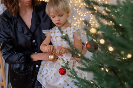 A mother with a 2-year-old daughter decorates the Christmas tree. Mom in a black suit, a girl in a white dress, her daughter hangs a red ball on the Christmas tree. Merry Christmas and New Year concept.