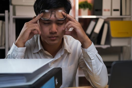 Exhausted young man with laptop in office working late sitting on desk in office overtime at night. Frustrated young businessman massaging his nose and keeping eyes closed while sitting at his working place in office.