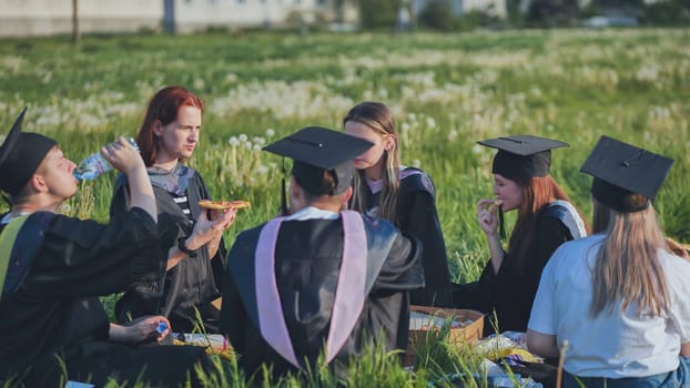 Graduates in black suits eating pizza in a city meadow