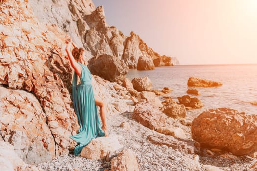 Side view a Young beautiful sensual woman in a red long dress posing on a rock high above the sea during sunrise. Girl on the nature on blue sky background. Fashion photo.