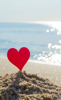 Plastic stick in shape of red heart in sand on sandy beach of sea shore on background of sea waves on sunny summer day close-up. Concept Love relationship romance amorousness amour St. Valentines Day