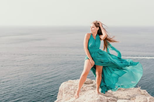 Side view a Young beautiful sensual woman in a mint long dress posing on a volcanic rock high above the sea during sunset. Girl on the nature on overcast sky background. Fashion photo