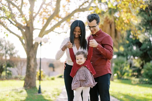 Smiling parents raising little girl by hands while standing in park. High quality photo