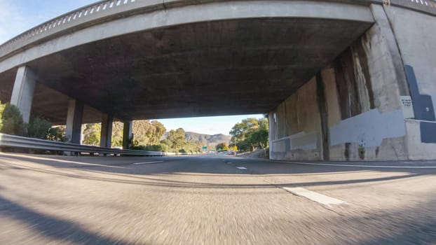 On a crisp winter day, a car cruises along the iconic Highway 1 near San Luis Obispo, California. The surrounding landscape is brownish and subdued, with rolling hills and patches of coastal vegetation flanking the winding road.