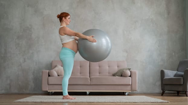 Pregnant red-haired woman doing arm exercises with fitball at home