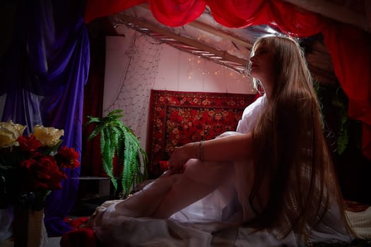 Beautiful European girl looking like Arab woman in red room with rich fabrics and carpets in sultan harem. Photo shoot of an oriental style odalisque. A model poses in sari as indian woman in india