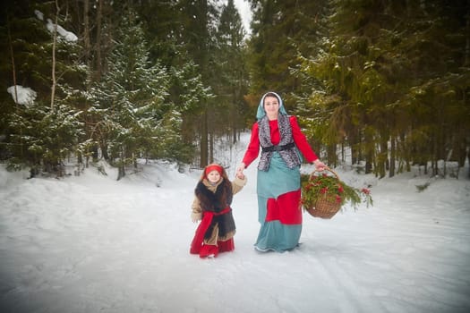 Family with mother and little daughter in stylized medieval peasant clothing in winter forest. The woman and child pose for fairy tale photoshoot in nature on a cold day. Concept of love, friendship