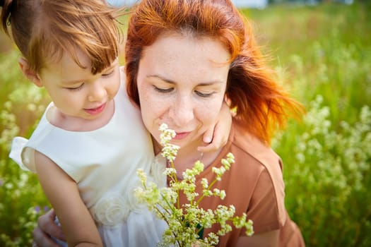 Happy female family with mother and daughter on green and yellow meadow full of grass and flower. Woman with red hair and blonde girl having fun, joy and hug in sunny summer day. Concept family love