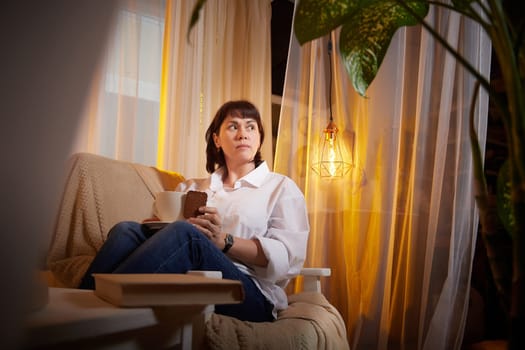 Nice girl with a mug of tea or coffee in cozy room. A middle-aged adult woman relaxing in the evening in the living room or bedroom in the evening