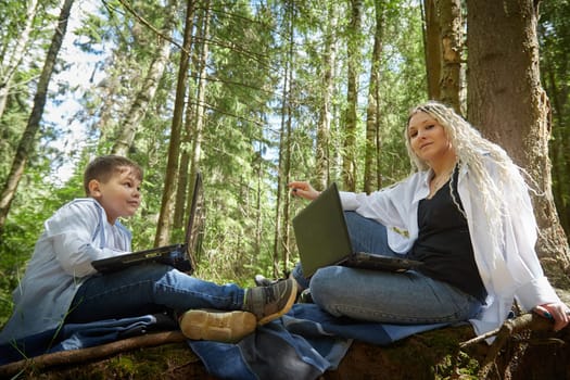 Mother and son with a laptops in the forest in summer. Fat young smart teenage boy and woman working with modern IT technologies in nature