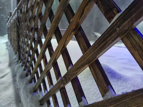 Wooden fence with rhombus planks and snowflakes on a winter evening with snow storm. Background, pattern, texture