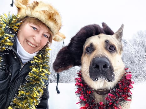 Funny Adult girl with shepherd dog in tinsel, fur caps taking selfie in a winter cold nature. Middle aged woman, big pet and Christmas or New Year