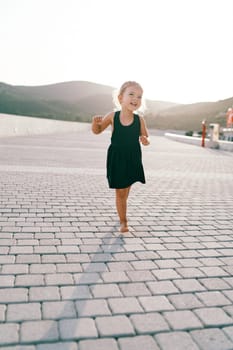 Little laughing girl walks barefoot on a tiled road, dancing and waving her arms. High quality photo