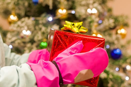 girl baby holds in pink gloves red christmas box before christmas tree.