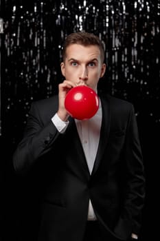 caucasian man in black jacket blowing red air balloon on glitter background. new year party