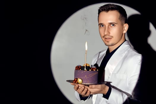 elegant caucasian man in white suit tuxedo holding cake with candle in the circle of light, copy space. Celebration