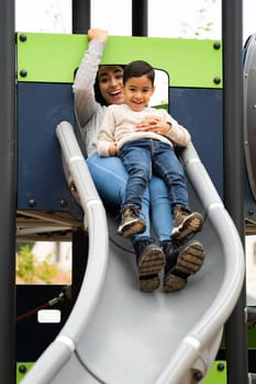Happy young latin mother having fun playing on the slide together in an outdoor playground with her son.