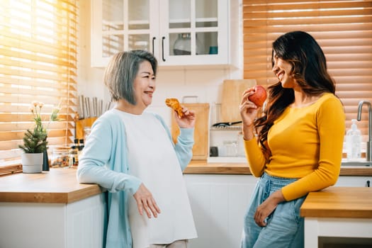 In the kitchen, an attractive young Asian woman and her mother share a moment of togetherness. Their smiles reflect the happiness and family bonding over food and learning.