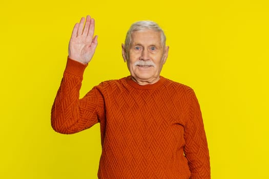 Old senior man smiling friendly at camera, waving hands gesturing hello greeting or goodbye welcoming with invitation hospitable expression. Elderly mature grandfather isolated on yellow background