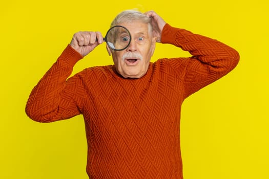 Investigator researcher scientist senior old man holding magnifying glass near face, looking into camera with big zoomed funny eye, searching, analyzing. Elderly grandfather on yellow background