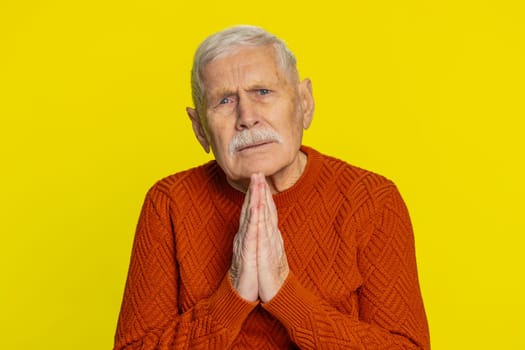 Please, God help. Senior old man praying, looking at camera and making wish, asking with hopeful imploring expression, begging apology, meditation. Mature grandfather isolated on yellow background
