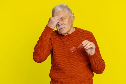 Exhausted tired senior old man takes off glasses feels eyes pain, being overwork burnout from long hours working. Sleepy exhausted elderly grandfather rubbing eyes isolated on studio yellow background