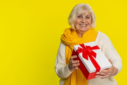 Positive smiling senior woman presenting birthday gift box stretches out hands, offer wrapped present career bonus, celebrating party, promotion discount sale. Elderly mature grandmother. Copy-space
