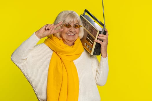 Senior old woman using retro tape record player to listen music, disco dancing favorite track, having fun, entertaining, fan of vintage technologies. Aged grandmother pensioner on yellow background