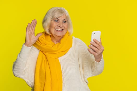 Happy senior old woman blogger influencer taking portrait selfie on smartphone, communicating record video call online with social media subscribers. Mature grandmother isolated on yellow background