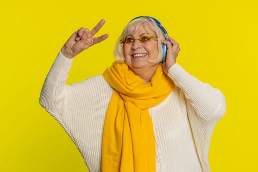 Excited senior stylish old woman listening music via headphones, dancing disco party fooling around having fun expressive gesticulating hands. Happy elderly grandmother pensioner on yellow background